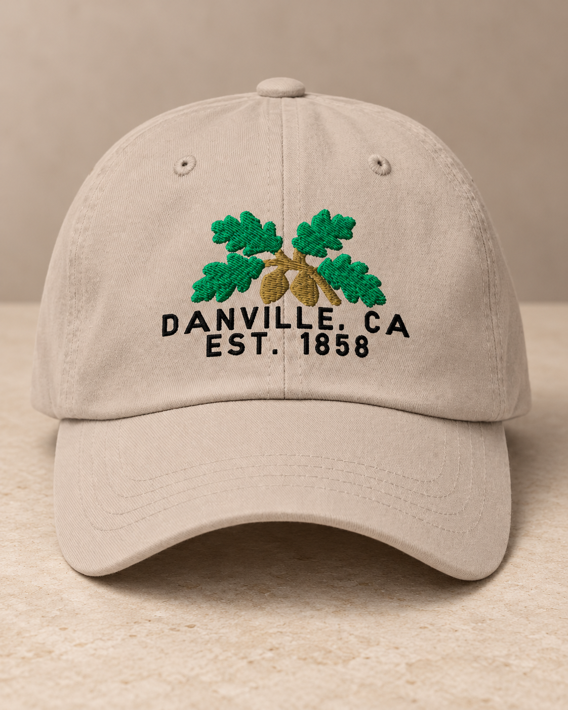 Digital studio presentation of the Stone Yupoong classic cap with green oak leaves, gold acorns and black Danville, CA / Est. 1858 embroidery on a warm stone surface.